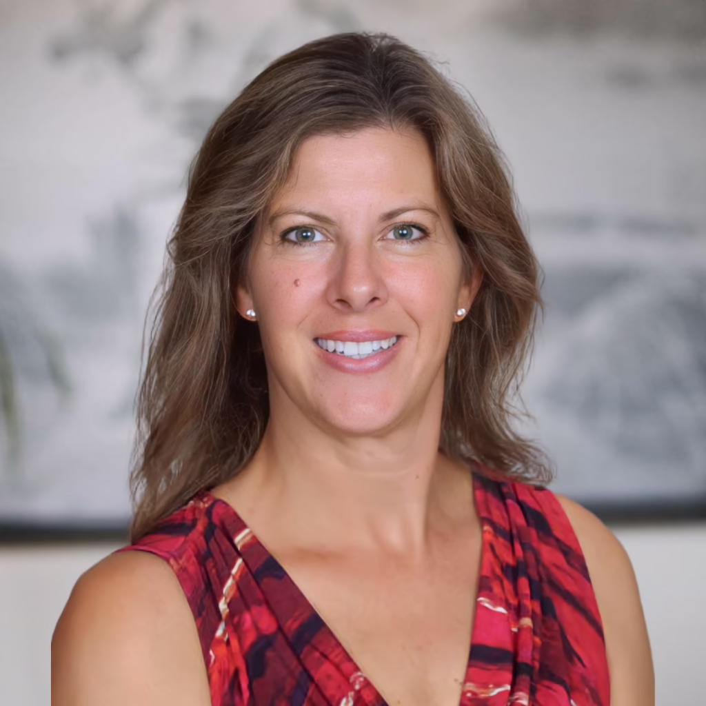 Professional headshot of a smiling woman with shoulder-length brown hair in a red patterned sleeveless top, blurred background