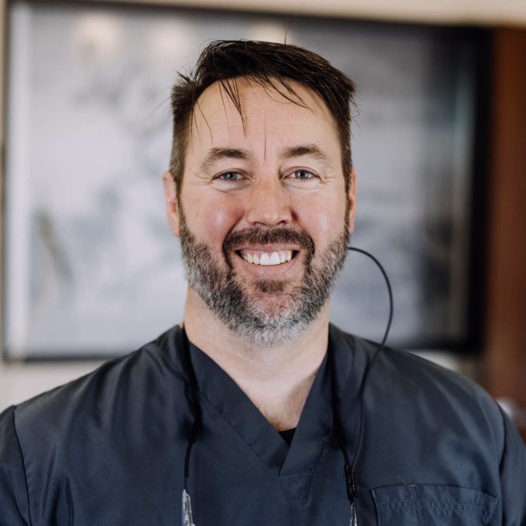 Smiling male healthcare professional in dark scrubs with a headset around his neck.