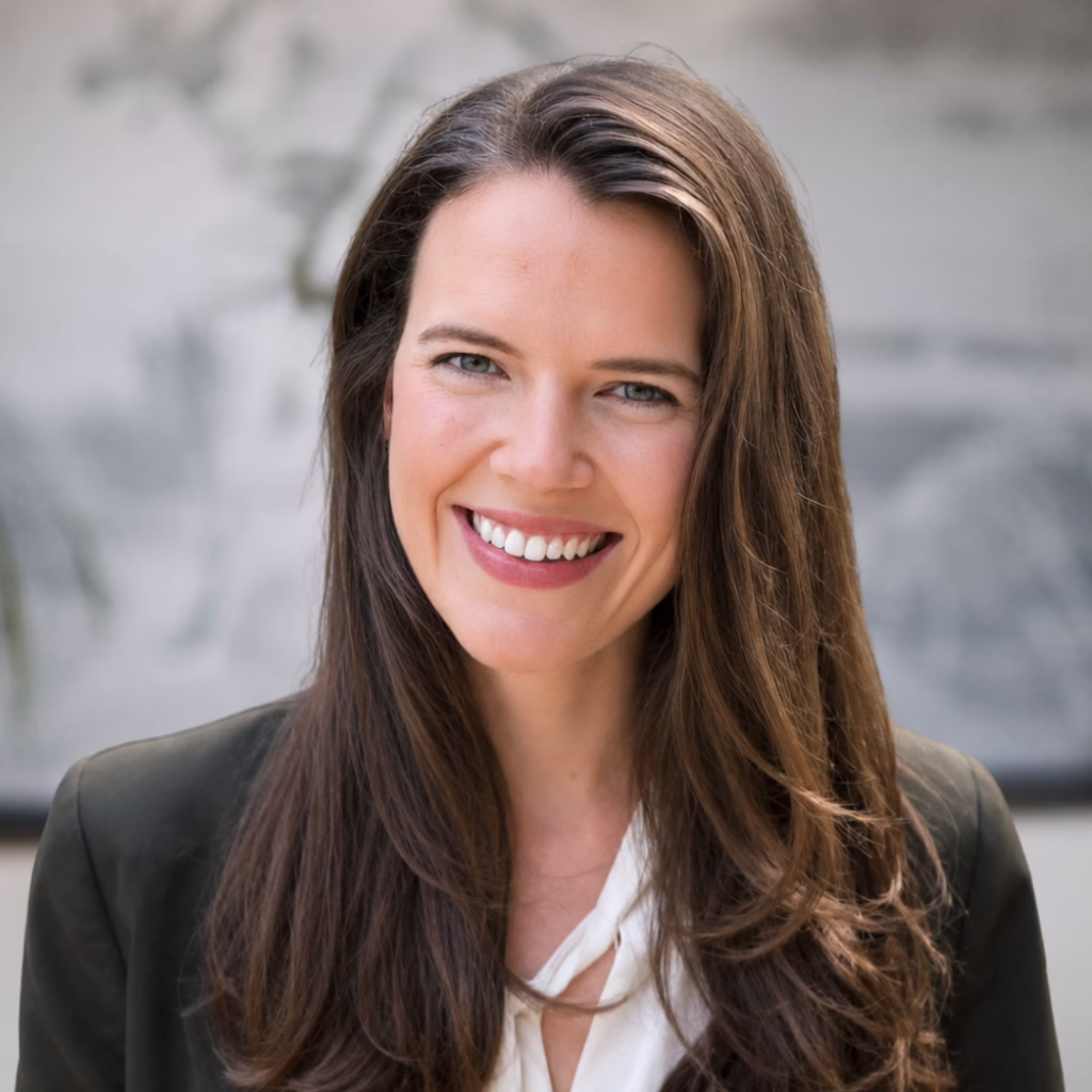 Professional headshot of a smiling woman with long brown hair, wearing a white blouse and dark blazer, neutral background.