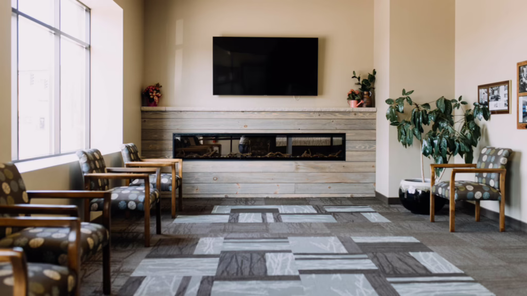 Waiting room lounge with beige walls, a long wooden fireplace mantel, a wall-mounted flat-screen TV, and potted plants on the mantle and right side.