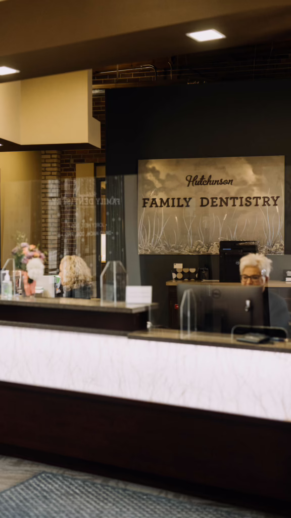 Reception desk at a family dentistry office with a sign reading Hutchinson Family Dentistry; staff member at a computer behind a clear divider.