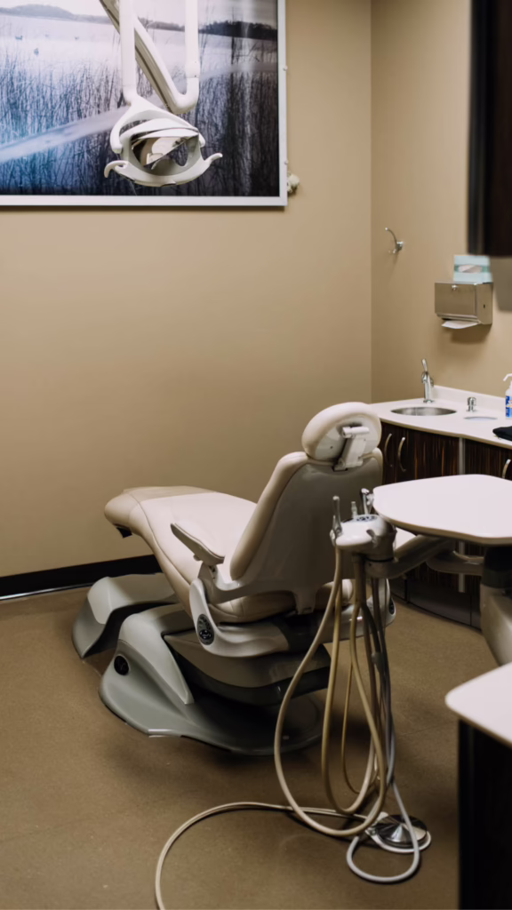 Dental chair and dental equipment in a beige-walled dental exam room, with a counter, sink, and framed artwork on the wall above.