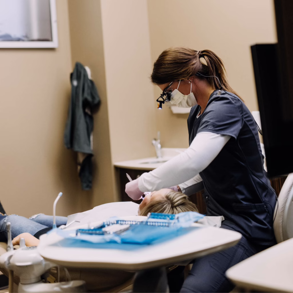Dental professional wearing mask and loupes treats a patient in a dental chair with dental tools laid out on the tray nearby