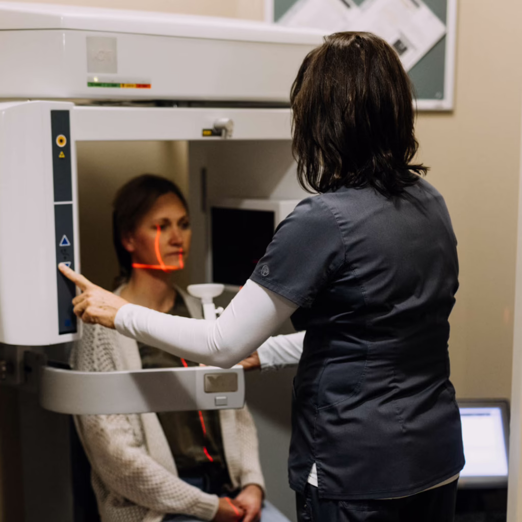 Healthcare professional adjusts an MRI machine while a patient sits in front, red alignment lasers visible for positioning.