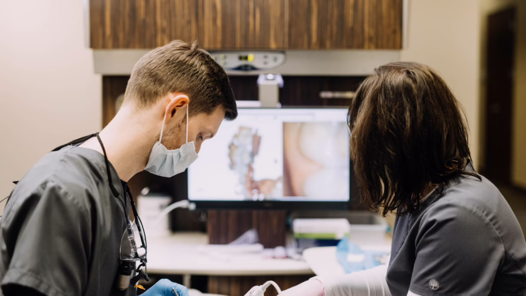 Two medical professionals wearing masks and scrubs perform a procedure in a clinic, with a monitor displaying medical images in the background.