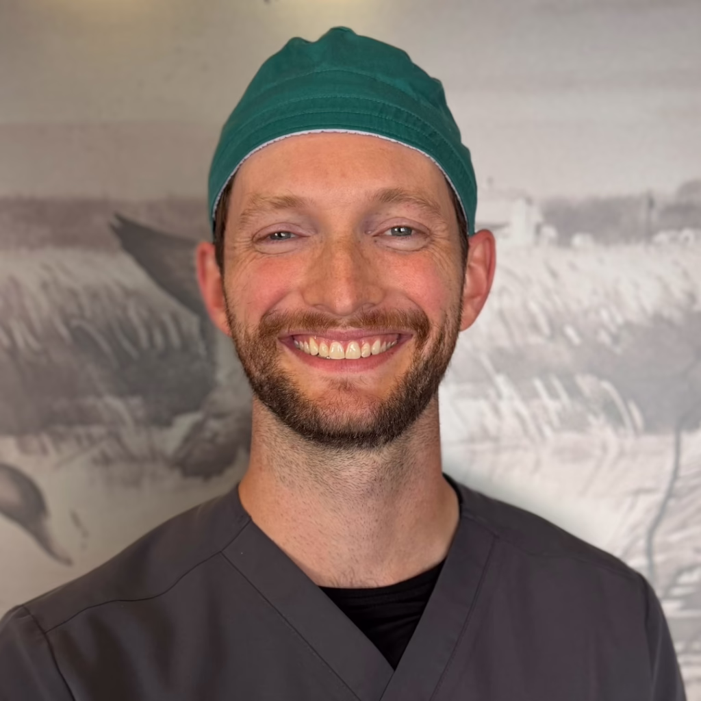 Smiling healthcare worker wearing gray scrubs and a green surgical cap.
